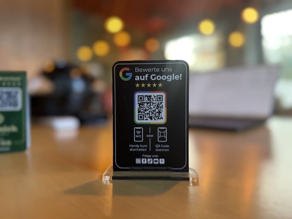 Ein kleiner schwarzer Aufsteller auf dem Standfuß auf einem Holztisch: „Bewerte uns auf Google!“ mit QR-Code, dazu die Hinweise Handy kurz dranhalten oder QR-Code scannen