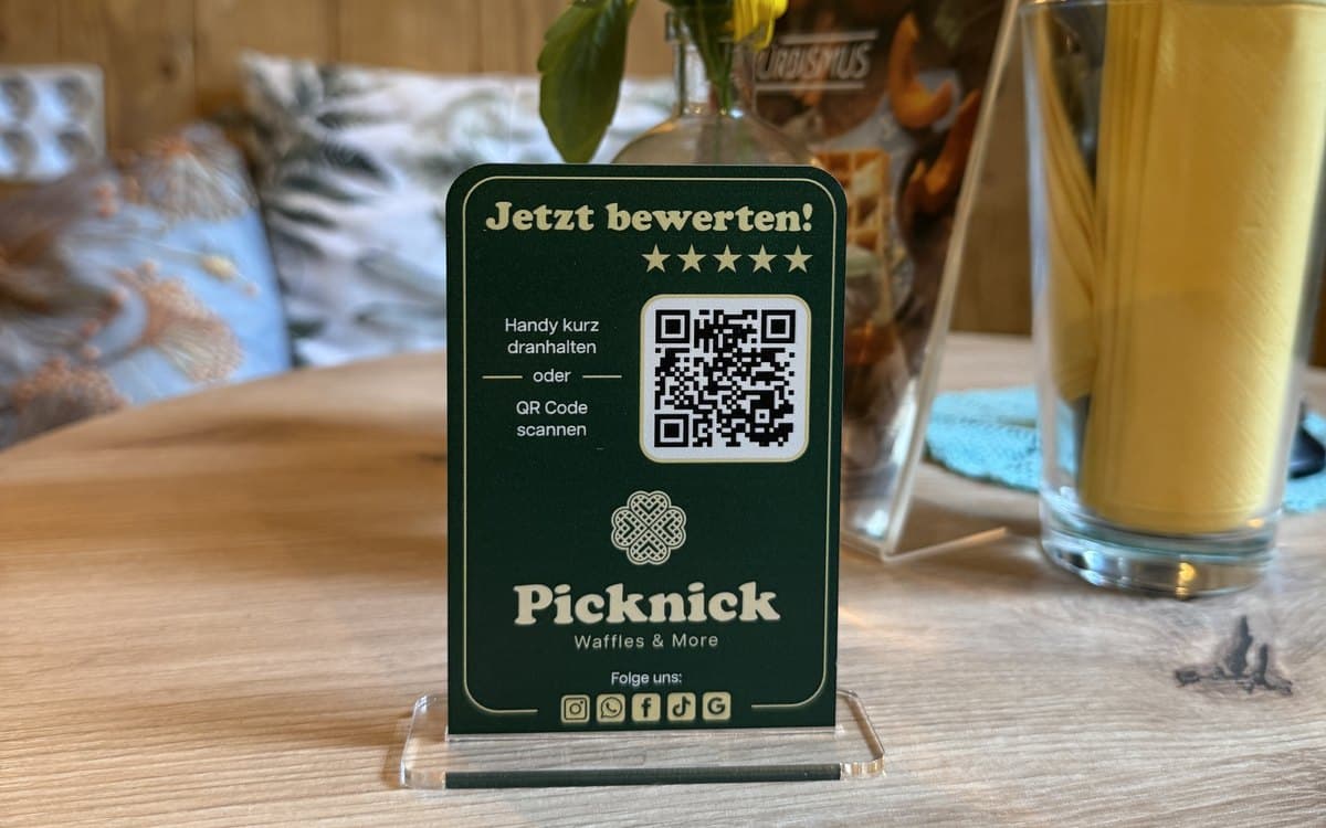 Die Rückseite des Aufstellers von Picknick Waffles and More auf einem Tisch: „Jetzt bewerten!“ mit fünf Sternen, QR-Code und NFC